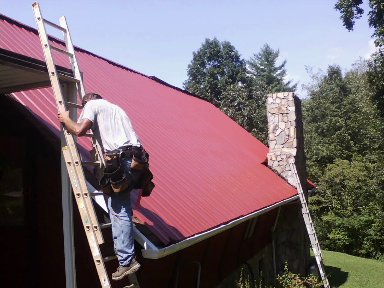 Roofer on a ladder beside a red metal roof and stone chimney — Soffit & Fascia Repair by Roofing Brothers in Avondale Estates